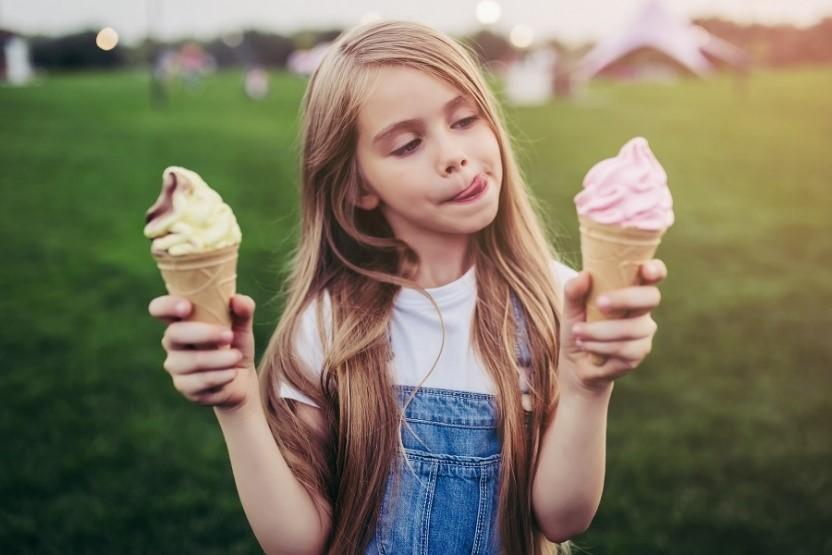 A young girl in a denim dress holding two ice cream cones, thoughtfully deciding which one to try first while standing in a park.