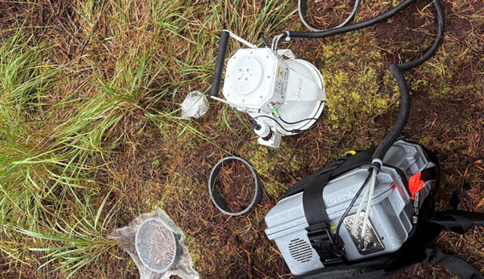 Photograph showing outdoor fieldwork equipment arranged on grassy, moss-covered ground, including a grey motor unit, white control component, long black hose and mesh scoop being used for surveying as part of the PEATFor Project
