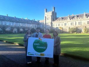 A photo of four people from the project team standing in front of the UCC quad holding a board with the EMIT-CHANGE and partner logos. Names in caption