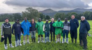 A group of people standing in a grassy field, some have protective boots. Hills and cloudy skies in the background.